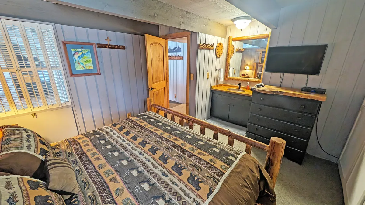 A serene bedroom with a bed dressed in patterned bedding, a wooden dresser with a mirror, TV, and subtle wall art. Natural light filters through the windows, creating a calm space ideal for relaxation and restful nights.