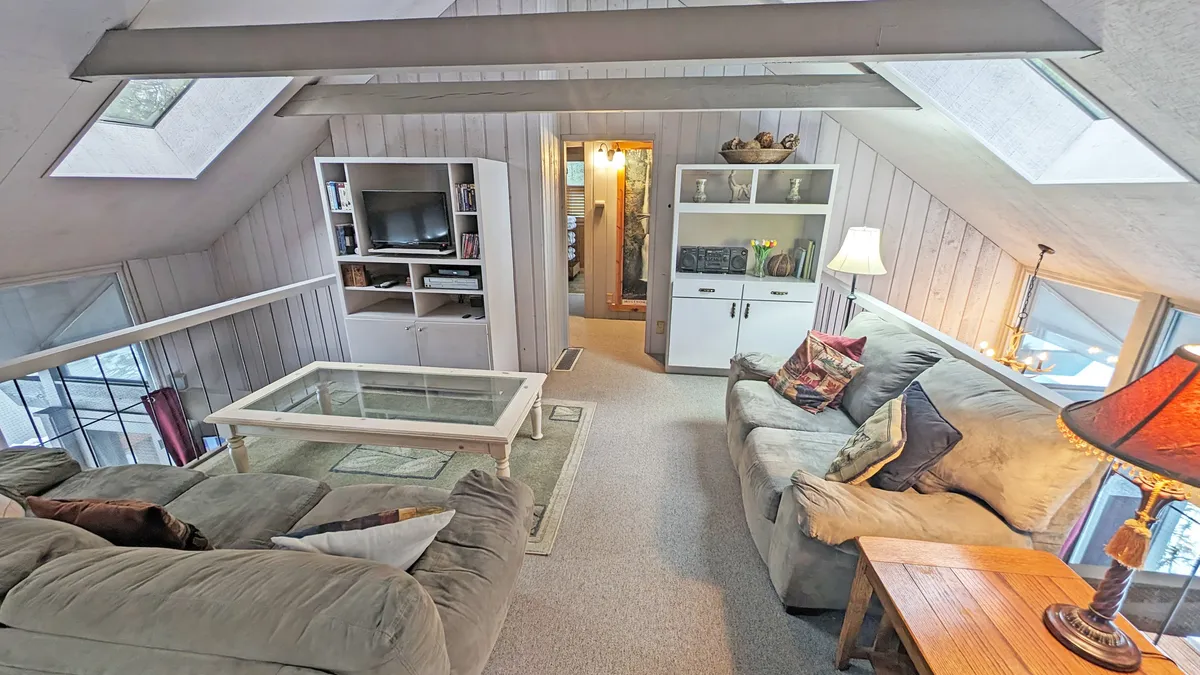 This image features a large sectional sofa arranged around a glass coffee table, with a bookshelf filled with decor and books along one wall. Skylights overhead flood the room with natural light, creating a warm, bright atmosphere ideal for relaxation and social activities.