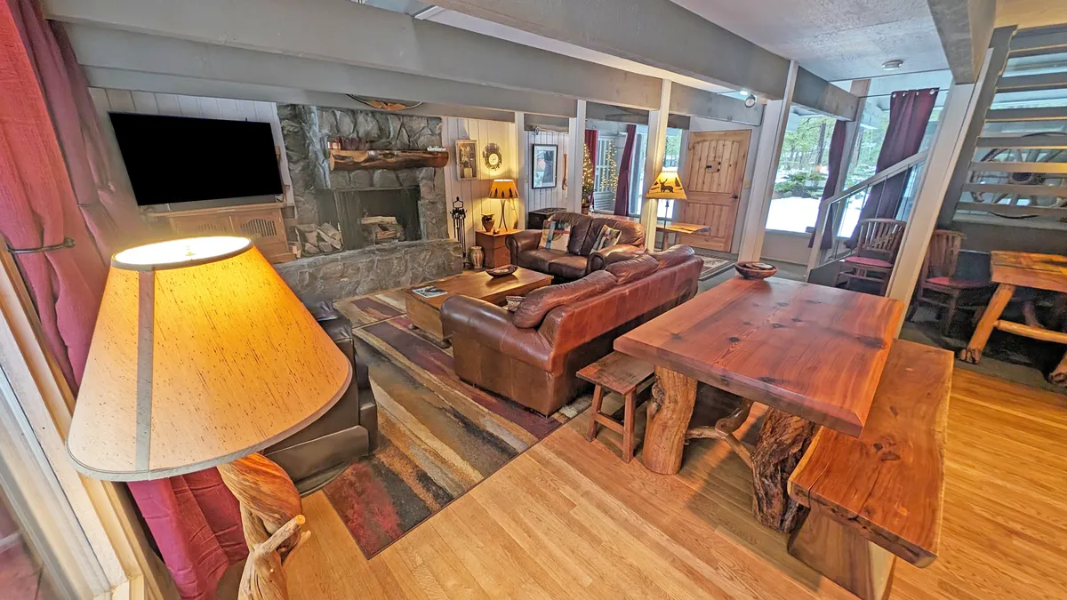The image showcases a warm, inviting interior with a plush leather sofa centered around a beautiful stone fireplace, and a spacious wooden dining table set for memorable group meals. The cozy ambiance is perfect for family gatherings, relaxing by the fire, and enjoying time together after Sunriver adventures.