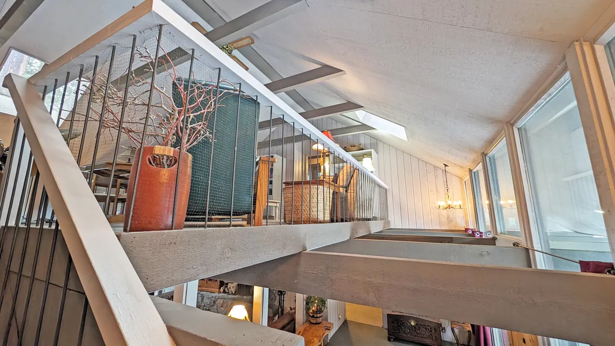 This photo showcases a loft area highlighted by exposed wooden beams and a balcony railing that overlooks the lower floor. Decorative plants and thoughtfully arranged furniture add a cozy, inviting vibe, and the abundant sunlight streaming in emphasizes the brightness of the space.