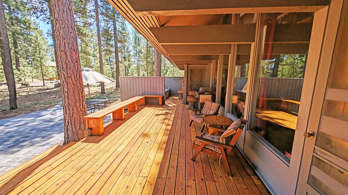 This image shows a wooden deck surrounded by tall trees, featuring comfortable outdoor seating and large windows looking into the home. The tranquil, natural setting is inviting for relaxing, socializing, or reading, offering a direct connection to the outdoors and the Sunriver environment.