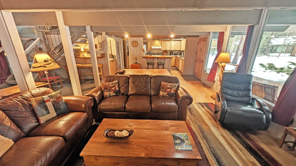 An open-concept living space that seamlessly integrates leather sofas, a wooden coffee table, and a spacious kitchen area in the background. Large windows let in abundant natural light, while a floor lamp and soft furnishings create an inviting zone for socializing or relaxing.