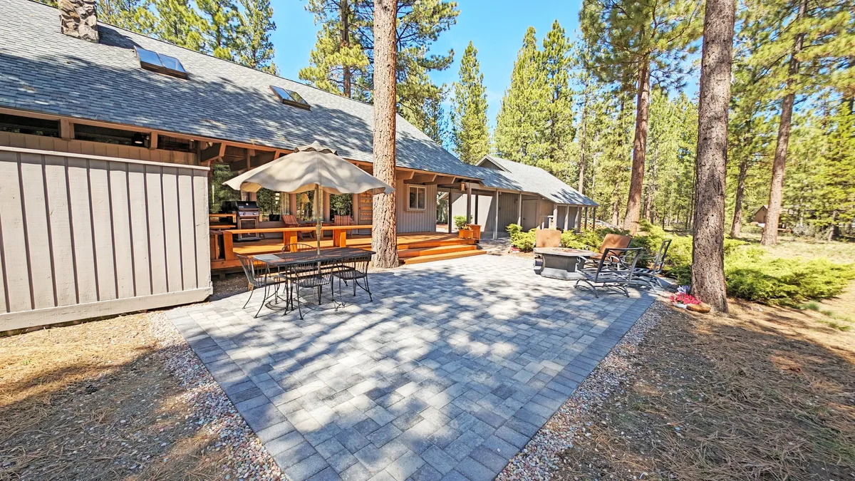A patio seating area with an umbrella and outdoor dining table, set against a backdrop of mature trees. The space is designed for al fresco meals, group gatherings, and cozy social moments in nature.