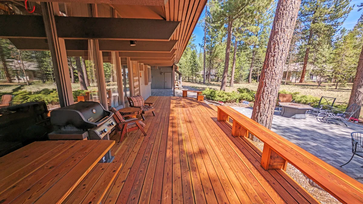 A spacious wooden deck surrounded by tall pine trees, featuring a gas grill and patio furniture. The deck exudes a relaxed ambiance, ideal for outdoor dining and gathering with family or friends. The grill is prominently positioned for easy access, while the seating area invites guests to linger and enjoy the natural setting.