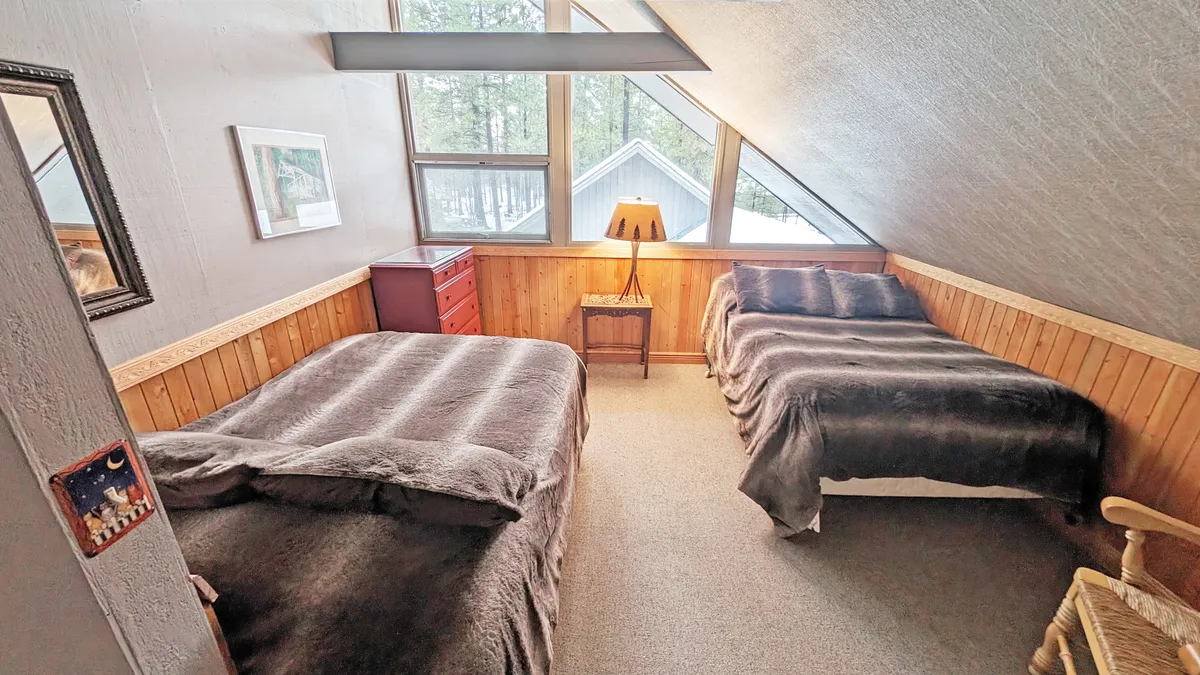 A cozy bedroom featuring two beds, a red bedside table, and a lamp set against warm-toned walls. The room is bathed in natural light from the windows, inviting a sense of comfort and relaxation, ideal for kids or cousins sharing a space.