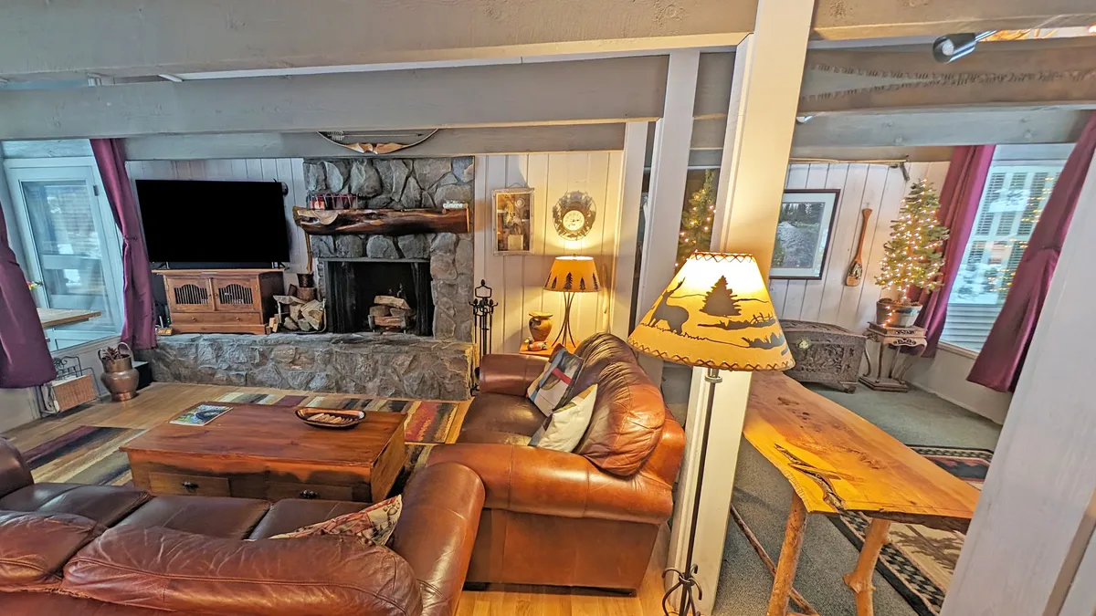 A welcoming living room featuring plush leather sofas arranged around a rustic stone-faced wood-burning fireplace. Warm accent lighting, a wooden coffee table, and a tastefully decorated Christmas tree enhance the cozy, communal atmosphere, with inviting textures and wood accents throughout.