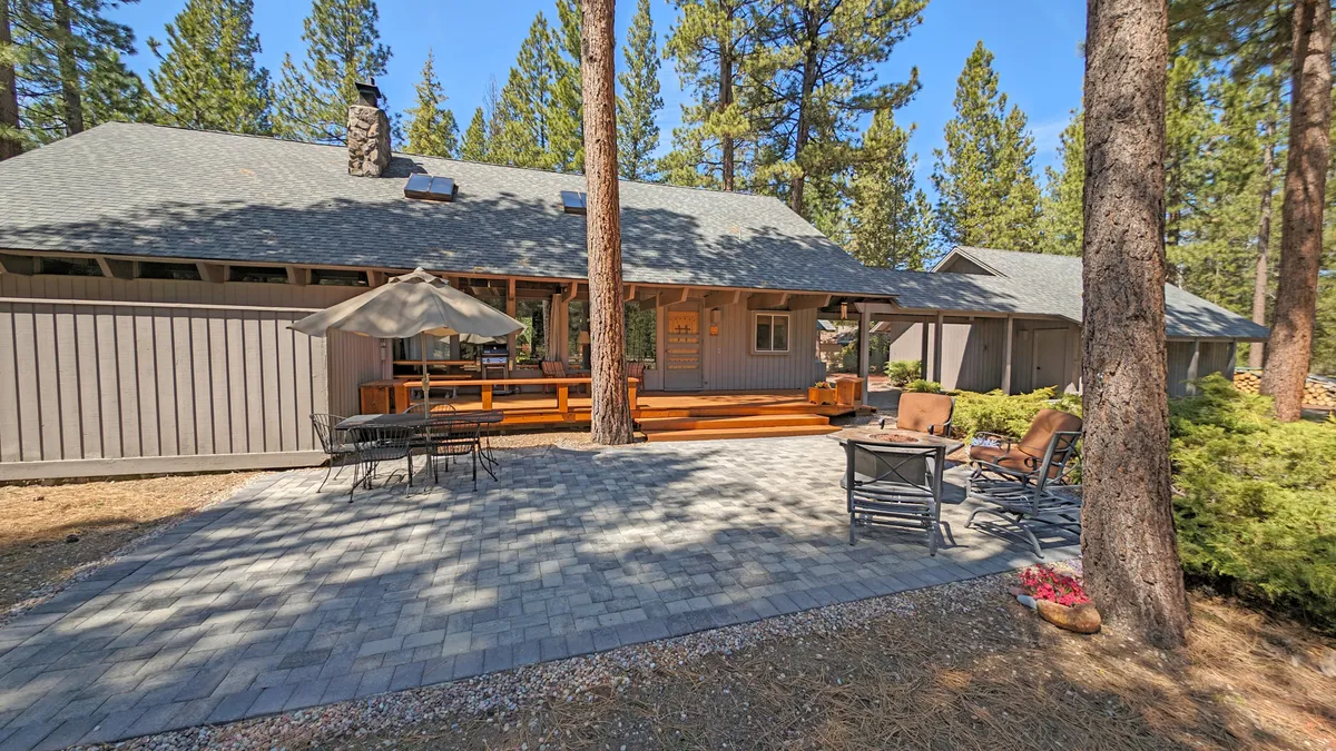 A spacious wooden deck with comfortable outdoor seating, a prominent stone chimney, and tall trees surrounding the area. The scene captures a sense of relaxed togetherness, with ample room for family and friends to gather outdoors.